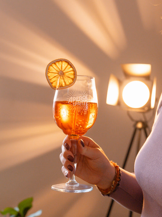Hand holding a glass of Aperol Spritz Candle with a slice of dried orange against a warm, blurred background